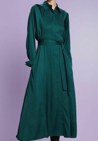 Robe en satin vert avec un col à revers, des manches longues, une taille nouée et des poches latérales, tombant en dessous du genou avec une texture lisse.
