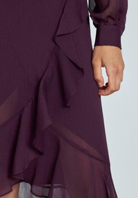 Robe violette avec des accents volantés ; réalisée en tissu léger et texturé ; présente des manches longues transparentes et un ourlet fluide.