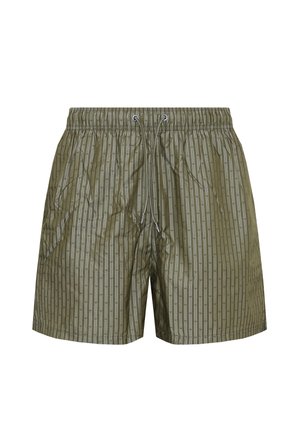 Shirtloze jonge man met tatoeages op zijn rechterarm en been, draagt groene gestreepte korte broek en zwarte slippers tegen een witte achtergrond.