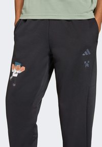 Černé joggingové kalhoty vyrobené z měkké tkaniny, s barevným grafickým motivem postavy z Minecraftu na jedné nohavici a detailem loga Adidas v tmavě šedé barvě.