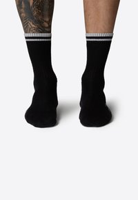 Calcetines negros acanalados con acentos de rayas blancas en el puño, con una textura suave y de longitud media hasta la pantorrilla. Tatuaje visible en la pierna.