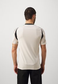 Světlá šedá sportovní tričko s černými akcenty, krátké rukávy, texturovaný vzor na ramenou a zaoblený lem. Hladký povrch látky.