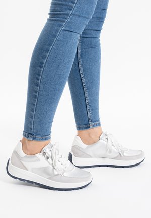 Silver and white athletic shoes with a textured upper, laces, and a navy sole, worn with blue skinny jeans.