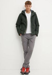 Jachetă verde cu glugă și fermoar, tricou alb, pantaloni gri cu buzunare și pantofi de drumeție roșii. Proiectată pentru activități în aer liber.