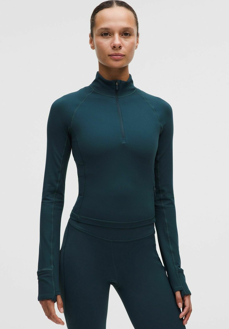 Kobieta w ciemnoniebieskiej, dopasowanej sportowej koszulce z długim rękawem i otworami na kciuki, w połączeniu z pasującymi leginsami o wysokim stanie, stoi na jednolitym tle.