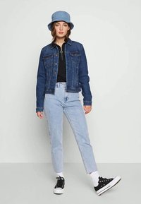 Mørkeblå denimjakke med knaplukninger og brystlommer, sammen med lyseblå jeans, en sort top og en lyseblå buckethat.