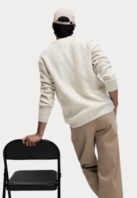 Ljusgrå ribbad tröja, beige byxor och en beige keps; lutad mot en svart hopfällbar stol med ena handen. Enkel och avslappnad design.