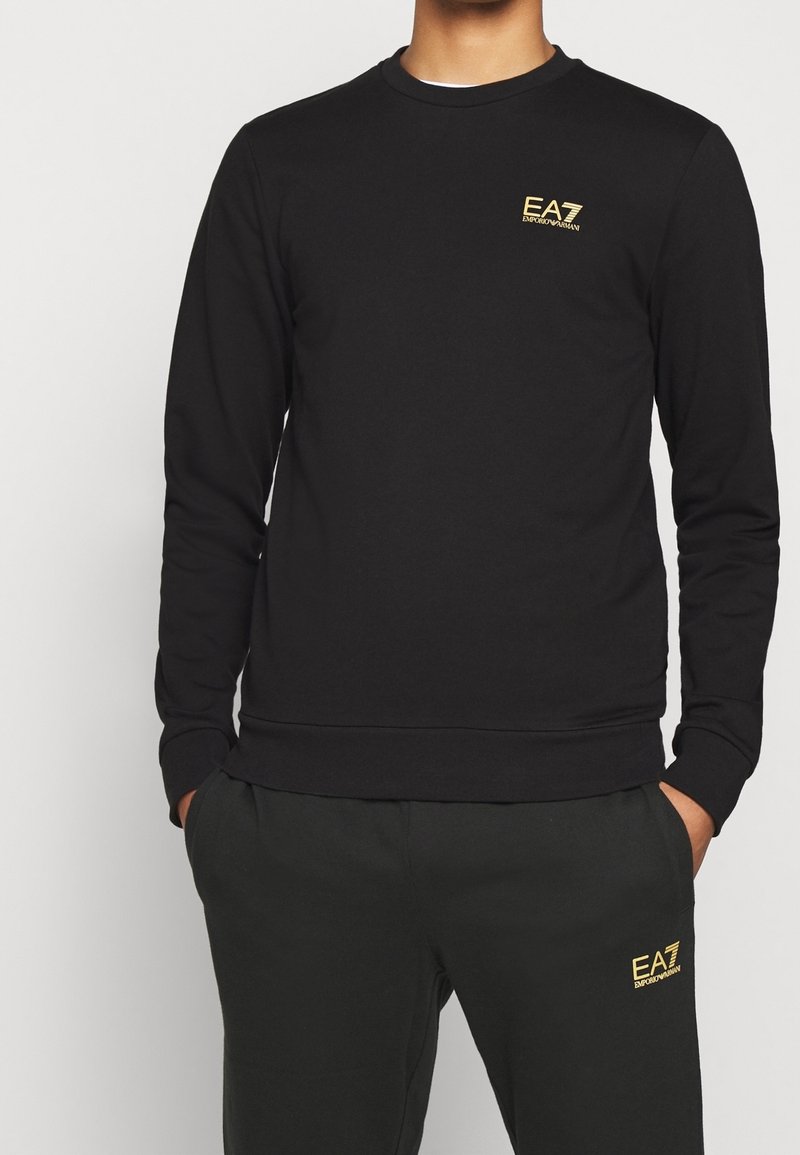 Hombre con sudadera negra de manga larga y pantalones a juego con los logotipos dorados de EA7 Emporio Armani en el pecho y el muslo. Manos en los bolsillos.
