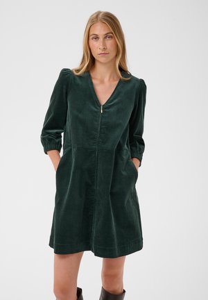 Femme aux cheveux blonds lisses portant une robe en velours côtelé vert foncé avec des manches bouffantes trois-quarts et les mains dans les poches.