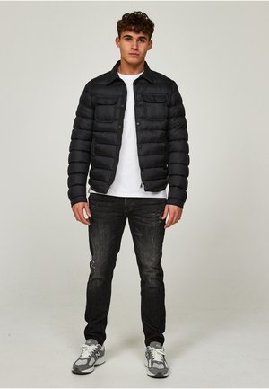 Jonge man staand, gekleed in een zwarte donsjas, wit T-shirt, zwarte spijkerbroek en grijze sneakers tegen een effen lichte achtergrond.
