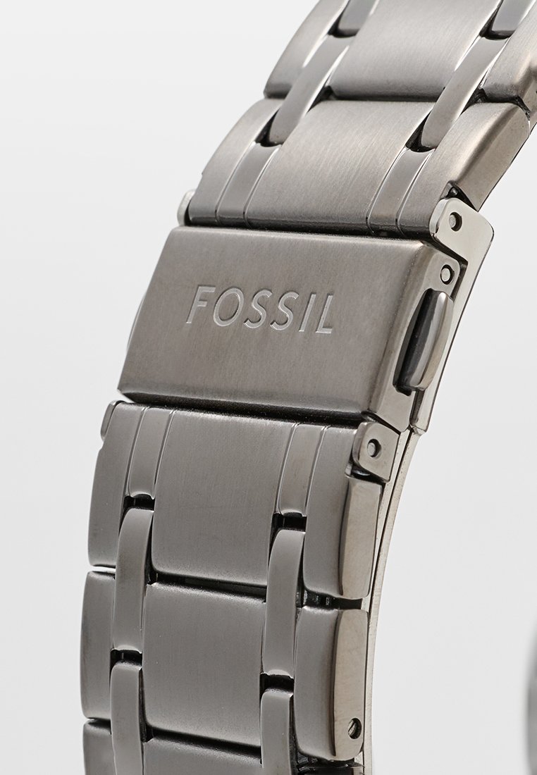 Banda de reloj de acero inoxidable con acabado cepillado, que presenta eslabones entrelazados, un cierre y el logo "FOSSIL" grabado en la parte ajustable.