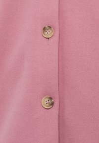 Ροζ μάλλινο ύφασμα με απαλή υφή, που διαθέτει δύο στρογγυλά, διαφανή κουμπιά με λουλουδένιο σχέδιο και εμφανείς ραφές.