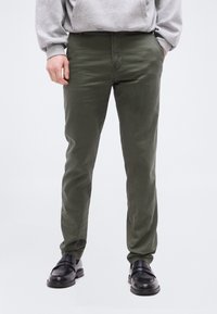 Ενήλικας που φοράει ελαφρώς εφαρμοστά χακί παντελόνια, ανοιχτό γκρι φούτερ και μαύρα δερμάτινα παπούτσια slip-on, στάσιμος με τα χέρια μερικώς στις τσέπες.