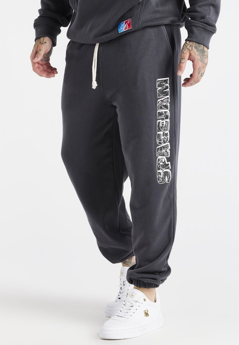 Man in zwarte trainingsbroek met witte trekkoord en verticale witte graphic op het been, gecombineerd met witte sneakers en een zwart sweatshirt.
