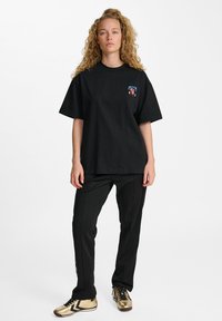 Zwarte katoenen T-shirt met ronde hals en korte mouwen, met een kleurrafische afbeelding op de linkerkant van de borst. Gepaard met zwarte broek en gouden schoenen.