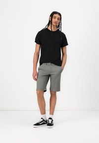 Tricou negru din bumbac, șorturi gri din bumbac și adidași negri cu talpă albă. Modelul stă cu o mână în buzunar, design minimalist.