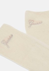 Cremige, plüschige Handschuhe mit weicher Textur; mit gesticktem "Guess" in glänzendem Silberfaden am Bündchen. Glatte, abgerundete Form.