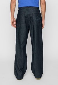 Mörk denimbyxor med avslappnad passform, försedd med dubbla bakfickor och subtila sömnadsdetaljer. Tyget har en slät textur och en stadig konstruktion.