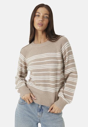 Vrouw met lang bruin haar die een beige trui met witte horizontale strepen en lichtblauwe spijkerbroek draagt, staand met handen bij de taille.