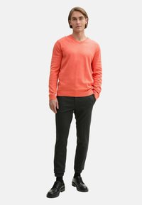 Pulover coral cu decolteu în V, dintr-un material moale de tricot, combinat cu pantaloni taperati gri închis și pantofi negri, stând în fața unui fundal alb simplu.