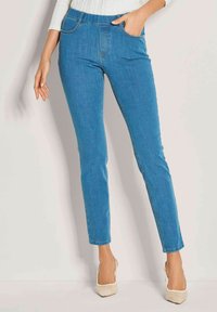 Frau in schlanken blauen Jeans und weißem strukturiertem Oberteil, eine Hand in der Tasche, stehend in beigen spitzen Absatzschuhen vor einer einfachen Wand.