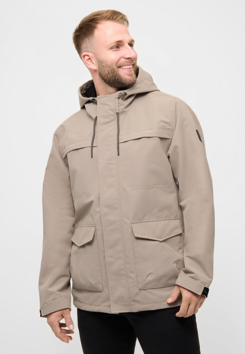 Smilende mand iført beige hættejakke til vinter med snore og forlommer med klap, ser mod højre mod en ensartet lys baggrund.