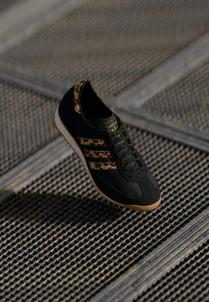 Sort sneakers med leopardmønstrede striber og kant, beige sål, placeret på en metalrist under blødt lys.