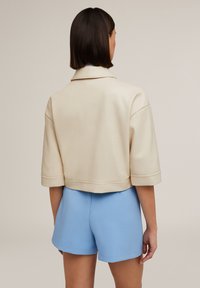 Giacca corta e cropped in finta pelle beige chiaro con colletto ampio e maniche arrotolate, abbinata a pantaloni corti blu.