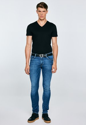 Noor mees seisab, seljas must V-kaelusega T-särk, sinised kitsad teksad musta vööga ja mustad tossud kummist tallaga, neutraalne taust.