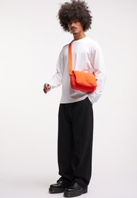 Borsa a tracolla arancione con design imbottito, indossata sopra una camicia bianca a maniche lunghe. Pantaloni neri larghi e scarpe nere completano l'outfit.