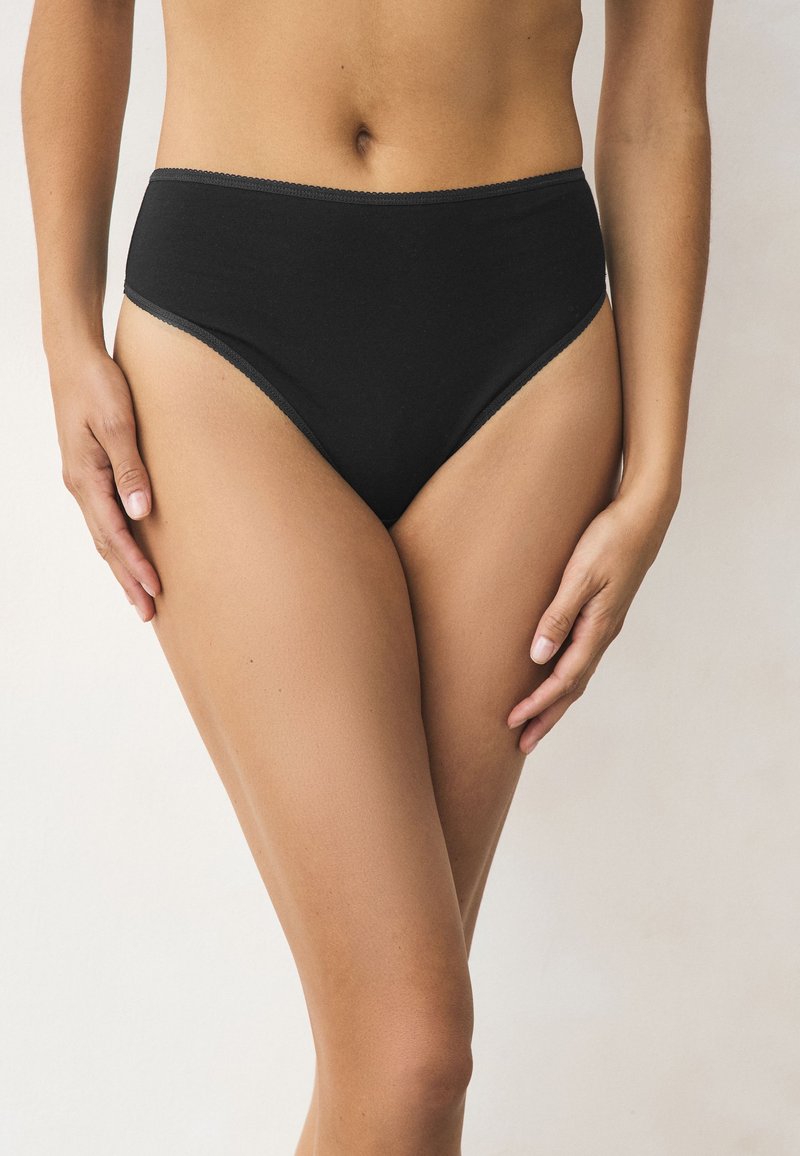 Mujer con ropa interior negra lisa de corte alto, de pie contra un fondo neutro, con las manos relajadas a los lados.