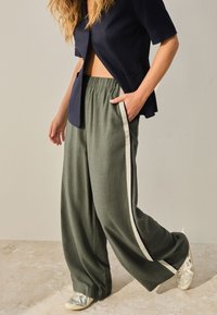Wide, olive green pants with a textured fabric, featuring a cream side stripe and elastic waistband. Paired with a dark blue short-sleeve top.