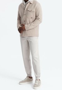 Hellbeige Jacke aus Wollmischung mit Kragen, zwei Brusttaschen, über einem grauen Hoodie getragen, kombiniert mit hellbeigen Jogginghosen und Turnschuhen.