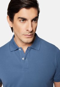 Męska koszulka polo w jednolitym niebieskim kolorze, z tradycyjnym kołnierzykiem, krótkimi rękawami i zapięciem na dwa guziki. Miękka tekstura.