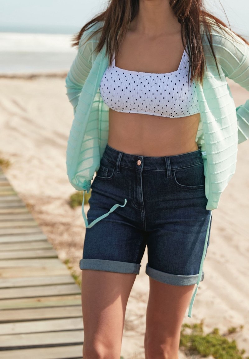 Jeune femme portant un crop top blanc à pois, un cardigan vert menthe et un short en jean foncé retroussé, debout sur un chemin en bois à la plage.