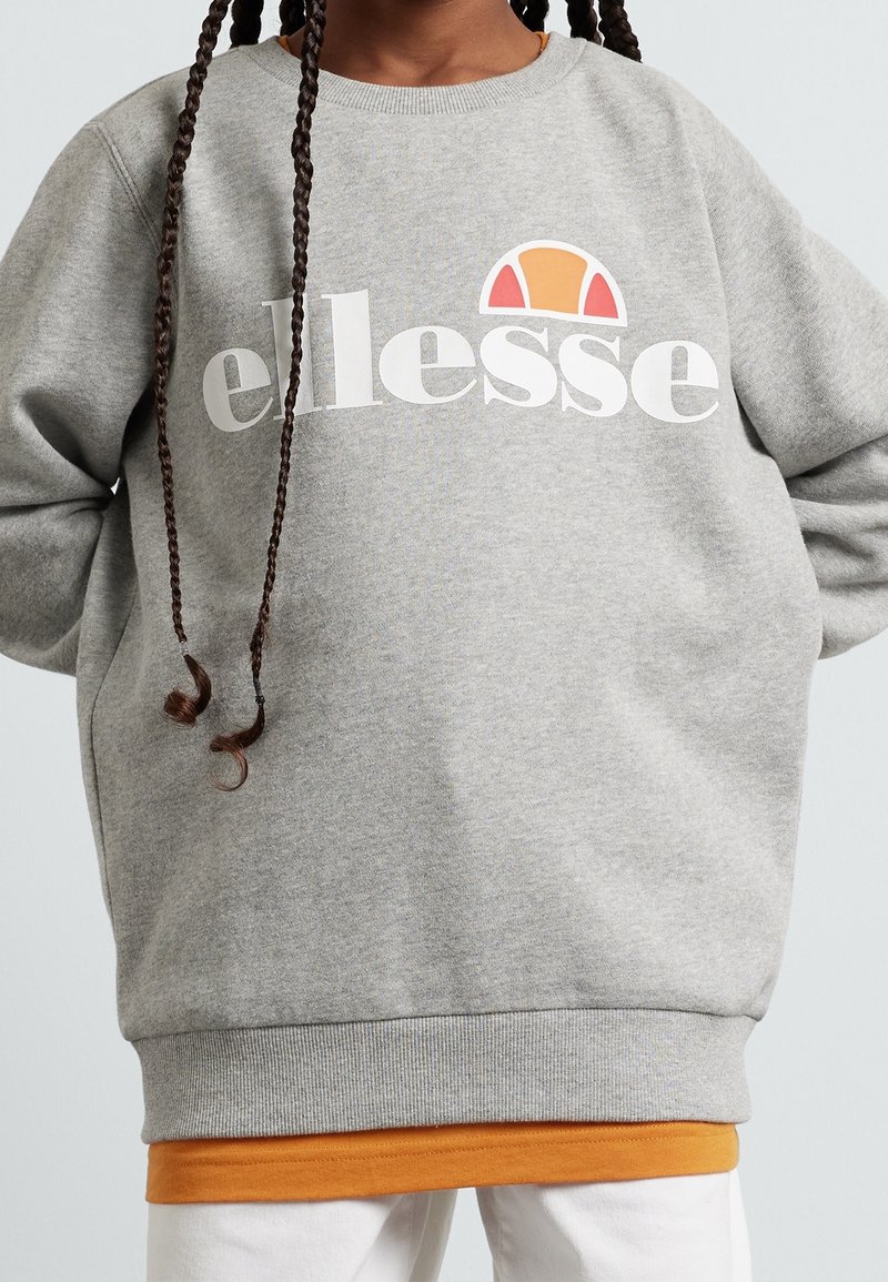 Grijze sweatshirt met wit "ellesse" logo, voorzien van een gekleurde halve cirkel grafiek. Geribbelde manchetten en zoom, gedragen over een oranje shirt.