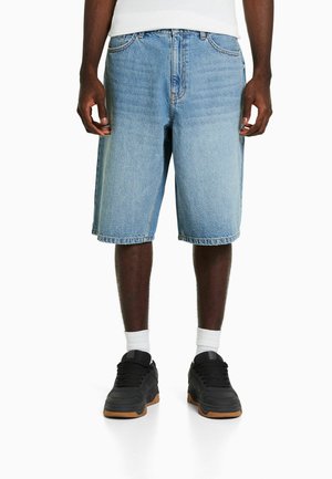 Shorts en jean bleu clair, longueur au genou, avec un design classique à cinq poches et une légère décoloration. Portés avec des baskets noires et des chaussettes blanches.