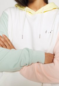 Bílá mikina s kapucí s akcenty v barvách mátová zelená, světle růžová a žlutá. Má malý stříbrný logo a měkkou, texturovanou látku.