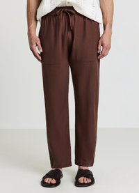 Braune, locker sitzende Hose mit elastischem Bund und zwei vorderen Taschen, aus strukturiertem Stoff. Getragen mit dunklen Sandalen.
