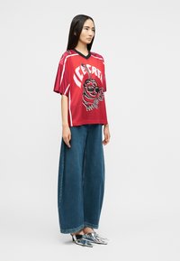 Γυναίκα με μακριά σκουρόχρωμα μαλλιά φοράει κόκκινη φαρδιά αθλητική μπλούζα με σχέδιο, μπλε παντελόνι με φαρδιά πόδια και ασημί μυτερά παπούτσια mule, στέκεται πλαγίως.