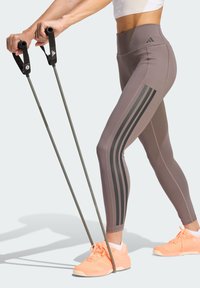 Γκρι κολάν με ψηλή μέση και μαύρες πλευρικές ρίγες, σε συνδυασμό με κοραλλί αθλητικά παπούτσια και μαύρες λάστιχες αντίστασης κρατημένες και στα δύο χέρια.