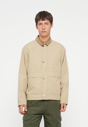 Beige jasje met een corduroy kraag, voorzien van een drukknoopsluiting aan de voorkant en twee grote voorzakken. Gelaagde structuur met een eenvoudig ontwerp.