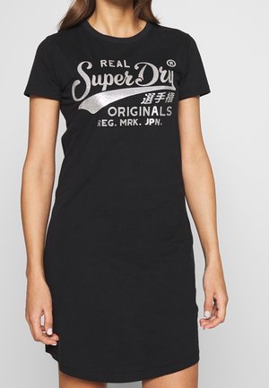 Zwarte katoenen jurk in t-shirtstijl met korte mouwen, voorzien van een zilveren "SuperDry" logo en Japanse tekst over de borst. Gladde textuur en een casual pasvorm.