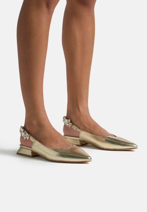 Scarpe slingback metalliche dorate con punta appuntita, tacco basso a blocco e decorazioni di cristallo sugli strap del tacco. Texture liscia.