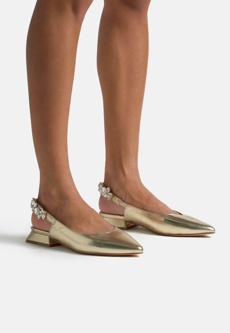 Scarpe slingback metalliche dorate con punta appuntita, tacco basso a blocco e decorazioni di cristallo sugli strap del tacco. Texture liscia.