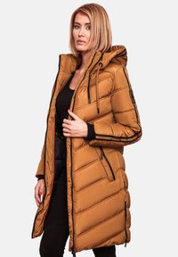 Long puffer jacket in tan, featuring a hood, diagonal stitching, zippered pockets, and black accents along the sleeves. Smooth, insulated fabric.