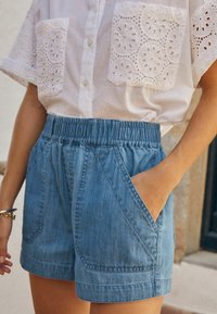 Biała bluzka zapinana na guziki z koronkowymi kieszeniami, zestawiona z wysokim stanem niebieskich szortów jeansowych z dużymi przednimi kieszeniami i elastycznym pasem.