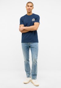 Granatowa bawełniana koszulka z okrągłym białym logo na lewej piersi, w połączeniu z jasnoniebieskimi dżinsami i białymi trampkami.