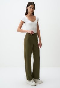 Weißes enges kurzärmliges Bodysuit kombiniert mit hoch taillierten olivgrünen Hosen. Das Model trägt weiße Sneaker. Glattes Material. Neutrale Hintergrund.