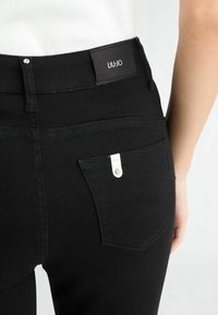Jean en denim noir avec une coupe slim, présentant une étiquette LIU-JO visible sur la ceinture et un petit accent métallique sur la poche arrière.